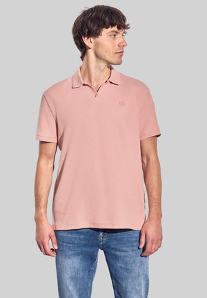 Camisa polo rosa claro con mangas cortas, cuello y un pequeño logo en el pecho. Combinada con unos jeans azules. Textura de tela suave.