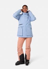 Helle blaue wasserdichte Jacke mit Kapuze, Fronttaschen und Reißverschlüssen. Kombiniert mit pfirsichfarbenen Hosen mit schwarzen Akzenten und schwarzen Skischuhen.