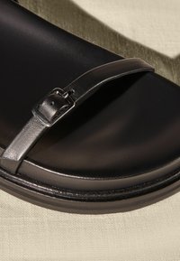 Svart sandal med en slät konstläderrem, ett rektangulärt metallspänne och en tjock, vadderad sula. Texturerad tygbakgrund.