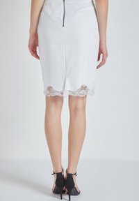 Mujer vestida con una falda blanca con borde de encaje y cierre en la parte trasera, combinada con zapatos negros de tacón alto con tira en el tobillo, de pie frente a un fondo blanco.