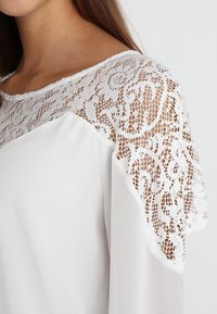 Blusa bianca con sovrapposizione di pizzo sulle spalle e un motivo floreale, corpo in tessuto liscio e design con scollatura ampia.