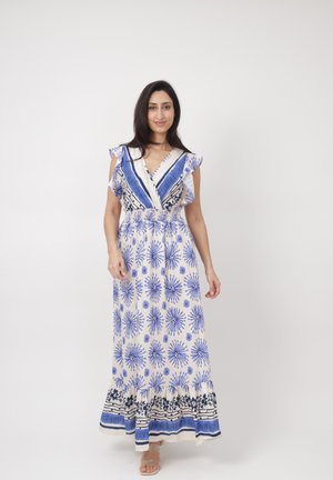 Femme debout portant une robe longue blanche sans manches avec des motifs floraux bleus et des rayures, souriante sur un fond blanc uni.