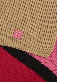 Écharpe tricotée avec une section côtelée beige et un patch logo rose, superposée sur un fond rayé rouge, noir et rose.