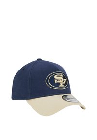 Marineblaue Kappe mit beigem Schirm, die an der Vorderseite ein gesticktes Logo "SF" in Gold und ein genähtes Paneeldesign aufweist.
