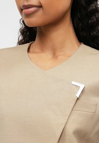 Bežs auduma tops ar V veida kaklu, ar sudraba adatu formas "7" un nelielu kabatas detaļu priekšpusē.