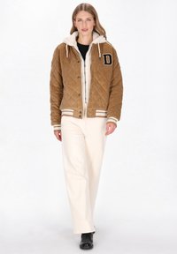 Ruskea korduroy-bomber-takki tikatulla kuviolla, norsunluunvärisellä hupullisella vuorella, logopohjaisella merkillä, resorikamppeilla hihansuissa ja helmassa; yhdistetty kermanvärisiin leveälahkeisiin housuihin.