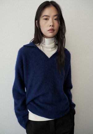 Joven con el pelo largo y oscuro, lleva un suéter azul sobre un jersey de cuello alto crema, de pie contra un fondo claro y liso.