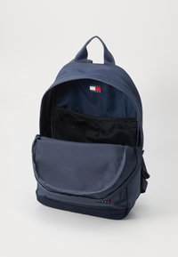 Sötétkék hátizsák tartós szövetből készült, nagy főrekesszel, elülső fedélzsebbel és belső hálós zsebbel rendelkezik. Tommy Jeans logó látható.