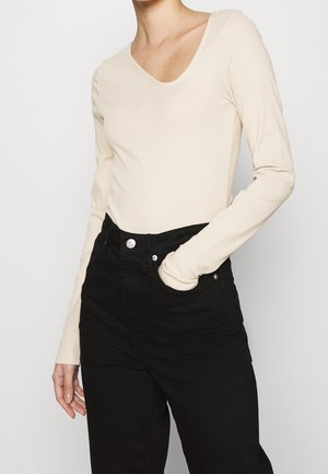 Persoon draagt een aansluitend crèmekleurig shirt met lange mouwen en een ronde hals, en een zwarte broek met hoge taille, één hand in de zak.