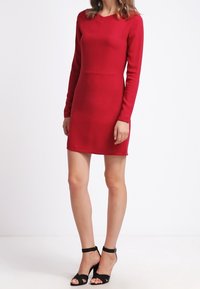 Punainen pitkähihainen mekko, jossa on hieman V-aukko, istuva bodice ja suora helma. Yhdistetty mustiin nilkkapantoihin. Sileä, joustava kangas.