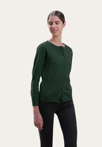 Cardigan vert à boutons avec des manches longues, un col rond, deux poches avant et une texture lisse. Porté par-dessus un jean foncé.