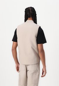 Beige ermeløs vest med rund hals, laget av bomull, sammen med en svart kortermet skjorte. Bakfra viser den en glatt tekstur og et rent design.