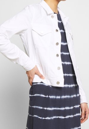Chaqueta de mezclilla blanca con botones plateados, bolsillos en el pecho y un corte corto, llevada sobre un vestido azul marino de teñido anudado con rayas horizontales.
