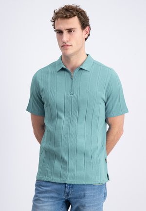 Man met krullend haar, die een teal textuur gebreid poloshirt met korte mouwen en rits draagt, staat met zijn handen achter zijn rug tegen een witte achtergrond.