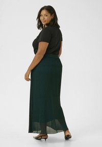 Camiseta negra y falda maxi plisada verde con cinturilla, capas transparentes y abertura. La modelo lleva zapatos de tacón negros y está de espaldas ligeramente.