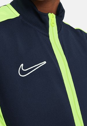 Marineblå jakke med lysegrønne accenter, har en høj krave og lynlås hele vejen foran. Nike-logo i hvidt på venstre brystområde.