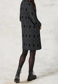Czarna sukienka w diamentowy wzór, z długimi rękawami i luźnym krojem. Noszona z czarnymi botkami za kostkę i czarnymi rajstopami. Teksturowana tkanina.