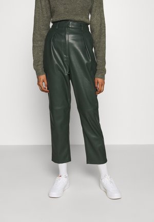 Personne portant un pantalon en cuir vert foncé taille haute, un pull en maille olive, des chaussettes blanches et des baskets blanches, debout devant un fond blanc.