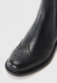 Stivale in pelle nera con dettagli brogue perforati, finitura liscia, punta arrotondata e suola testurizzata. Presenta un logo impresso sul lato.