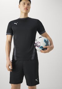 Svart atletisk t-shirt och shorts med grå detaljer, hållande en fotboll med svart och vit design och turkosa och rosa detaljer.