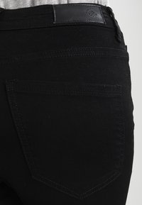 Pantalones de mezclilla negra con una textura suave, un bolsillo trasero y un parche de cuero en la cinturilla con un logo circular y detalles de costura.