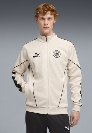 Jeune homme aux cheveux roux portant une veste zippée crème de Manchester City avec les logos noirs Puma et King, se tenant devant un fond gris.
