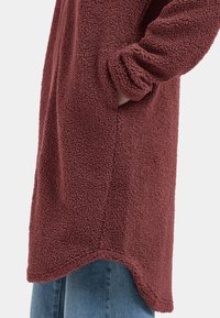 Bordeaux fleece jack met een gestructureerd oppervlak, voorzien van zijzakken, een gebogen onderkant en opgerolde manchetten, gedragen over een blauwe jeans.