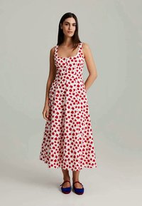 Witte midi-jurk met rood bloemenpatroon, vierkante halslijn, aansluitende lijfje, geplooide rok en een gladde textuur. Draagt met blauwe platte schoenen.