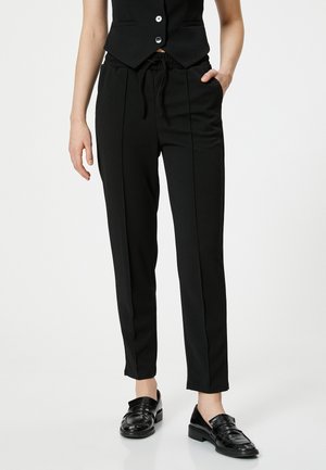 Pantalon noir fuselé avec une taille élastique et un cordon de serrage, doté de poches latérales et d'une finition lisse et texturée. Assorti à des mocassins noirs.