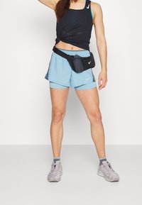 Musta hihaton toppi, vaaleansiniset shortsit, musta vyölaukku, jossa vesipullo, ja harmaat kuvioidut lenkkarit.