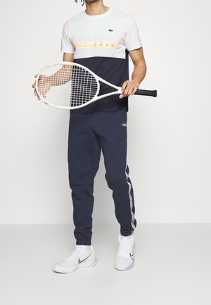 Completo sportivo composto da una maglietta a blocchi di colore bianca e blu navy con dettagli arancioni, pantaloni della tuta blu navy con strisce laterali e scarpe da tennis bianche.