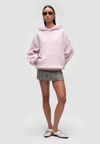 Felpa oversize rosa chiaro con tasca frontale, abbinata a una mini gonna grigia a fantasia e scarpe bianche con cinturini; modella che indossa occhiali da sole.