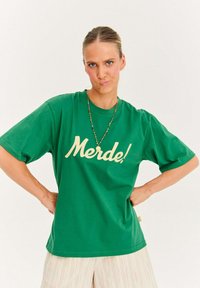 Zelené bavlnené tričko s krátkymi rukávmi, na prednej strane s veľkým krémovým nápisom "Merde!". Vo voľnom strihu s okrúhlym výstrihom.