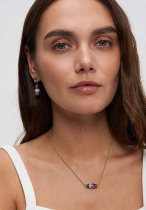 Young woman wearing a white top, gold chain necklace with multicolored beads, and matching gold drop earrings with colorful beads.