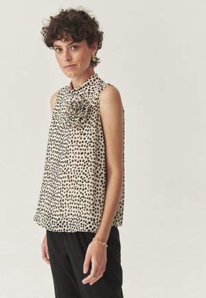 Femme avec des cheveux frisés courts portant un débardeur beige à pois avec des détails floraux, un pantalon noir et des bijoux dorés, sur fond uni.