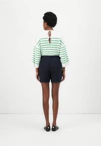 Langan Ärmelschnitt in Grün-weiß gestreift mit Rückenschnürung, kombiniert mit dunklen Shorts und schwarzen Slippern, von hinten aufgenommen.