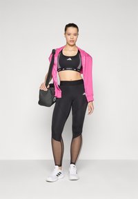 Black sports bra, pink hoodie, and black leggings with mesh cutouts. White sneakers with three black stripes; a black bag hangs from shoulder.