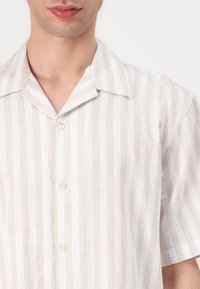 Ανοιχτόχρωμο κοντομάνικο πουκάμισο με κατακόρυφες μπεζ και λευκές ρίγες. Διαθέτει γιακά τύπου camp και δύο κουμπιά μπροστά.