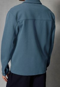 Lichtblauw overhemd met lange mouwen en een kraag, uitgevoerd in een textuurstof en voorzien van knoopmanchetten, gezien van de achterkant tegen een neutrale achtergrond.