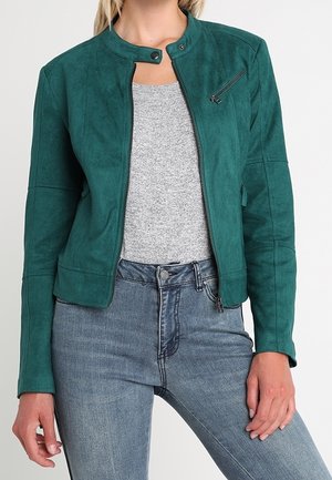 Frau trägt eine grüne Jacke mit Reißverschluss über einem grauen Hemd und hellblauen Jeans, steht mit entspannten Händen an den Seiten.