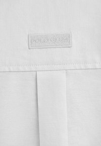 Tessuto bianco con una finitura texturizzata, caratterizzato da un'etichetta cucita che riporta la scritta "POLO GLUE". Design semplice con una superficie liscia e senza pattern aggiuntivi.
