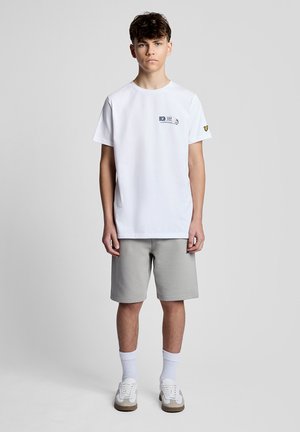 Teenagedreng står oprejst iført en hvid T-shirt med lille logo, grå knælange shorts, hvide sokker og hvide sneakers på en ensfarvet baggrund.