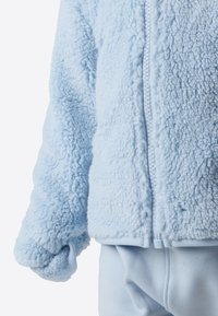 Jachetă bleu deschis din fleece cu suprafață texturată și pufoasă, prevăzută cu fermoar frontal și mâneci cu manșete. Asortată cu pantaloni în aceeași nuanță de bleu deschis.