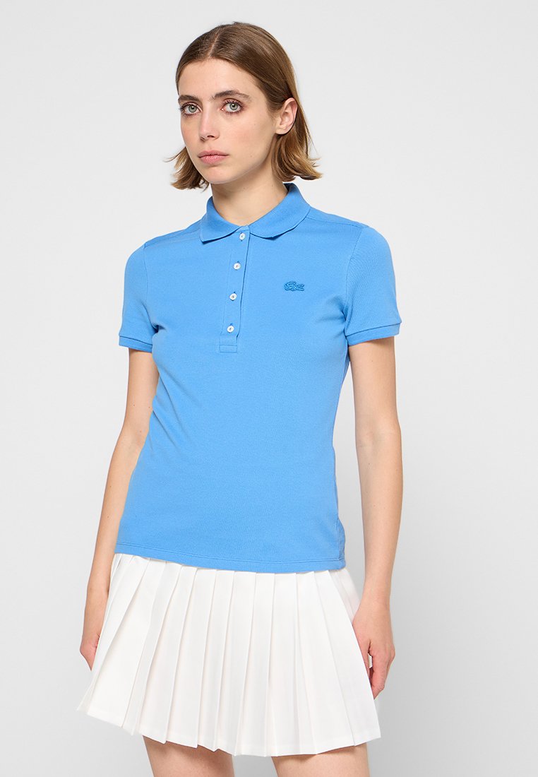 Polo shirt bleu avec un col côtelé et des manches courtes. Présente une patte de boutonnage à trois boutons et un logo brodé. Porté avec une jupe plissée blanche.