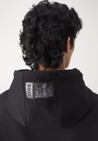 Homme aux cheveux noirs bouclés portant un sweat à capuche noir avec un écusson "BOSS Super Bowl" au dos de la capuche.
