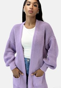 Lila stickad cardigan med lös passform, öppen framsida, ribbad textur och två framfickor. Buren över en vit åtsittande topp och denim.