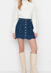 Denim rok met een golvende rand, voorzien van een sluiting met knopen aan de voorkant. Gecombineerd met een crème coltrui en beige kniehoge laarzen.