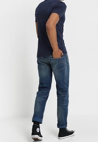 Tmavě modré tričko s krátkým rukávem, slim-fit, v kombinaci s ohrnutými modrými džíny a černými vysokými teniskami s bílou podrážkou, pohled zezadu.