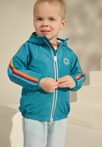 Türkiz kapucnis dzseki elülső cipzárral, a kezeknél piros, narancs és kék csíkokkal díszítve, valamint logóval a mellkason.