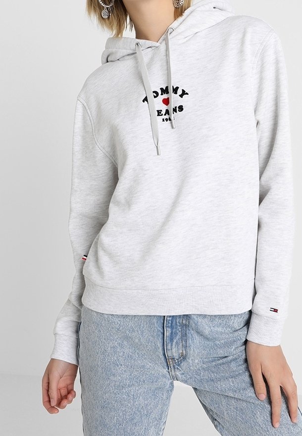 Sweat à capuche gris clair avec cordon, arborant le logo "TOMMY JEANS" en noir et rouge. Poignets et ourlet côtelés, porté avec un jean bleu clair.